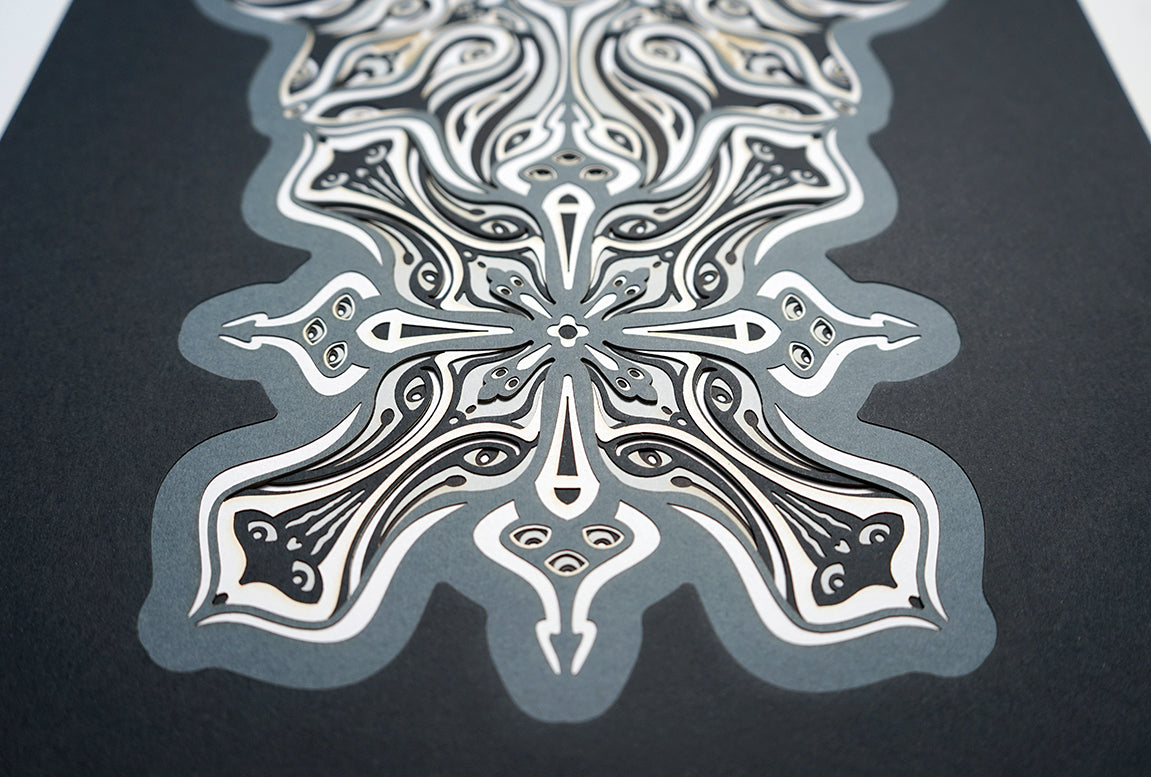 Monochrome layered paper artwork titled Metamorphs, made of intricate geometric mandalas that reveal totemic faces.