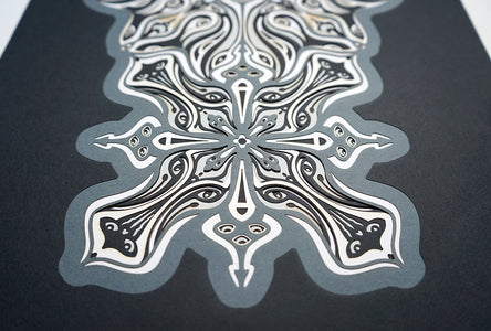 Monochrome layered paper artwork titled Metamorphs, made of intricate geometric mandalas that reveal totemic faces.
