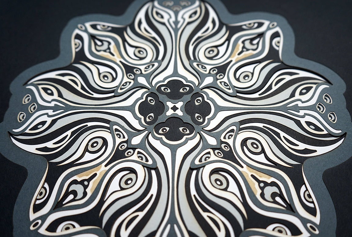 Monochrome layered paper artwork titled Metamorphs, made of intricate geometric mandalas that reveal totemic faces.