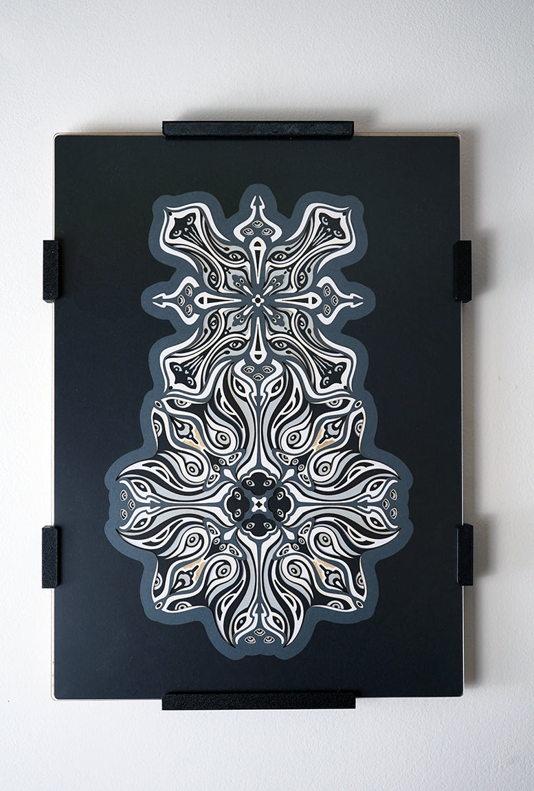 Monochrome layered paper artwork titled Metamorphs, made of intricate geometric mandalas that reveal totemic faces.