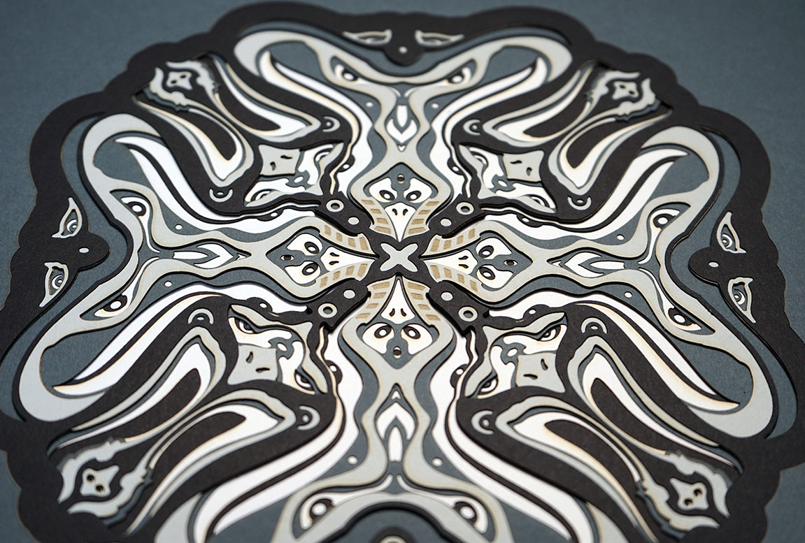 Monochrome layered paper artwork titled Metamorphs, made of intricate geometric mandalas that reveal totemic faces.