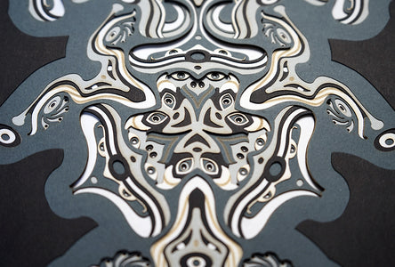 Intricate monochrome paper layers arranged in a mandala structure, creating emerging spirit faces and abstract creatures.