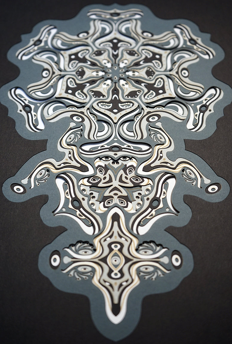 Intricate monochrome paper layers arranged in a mandala structure, creating emerging spirit faces and abstract creatures.