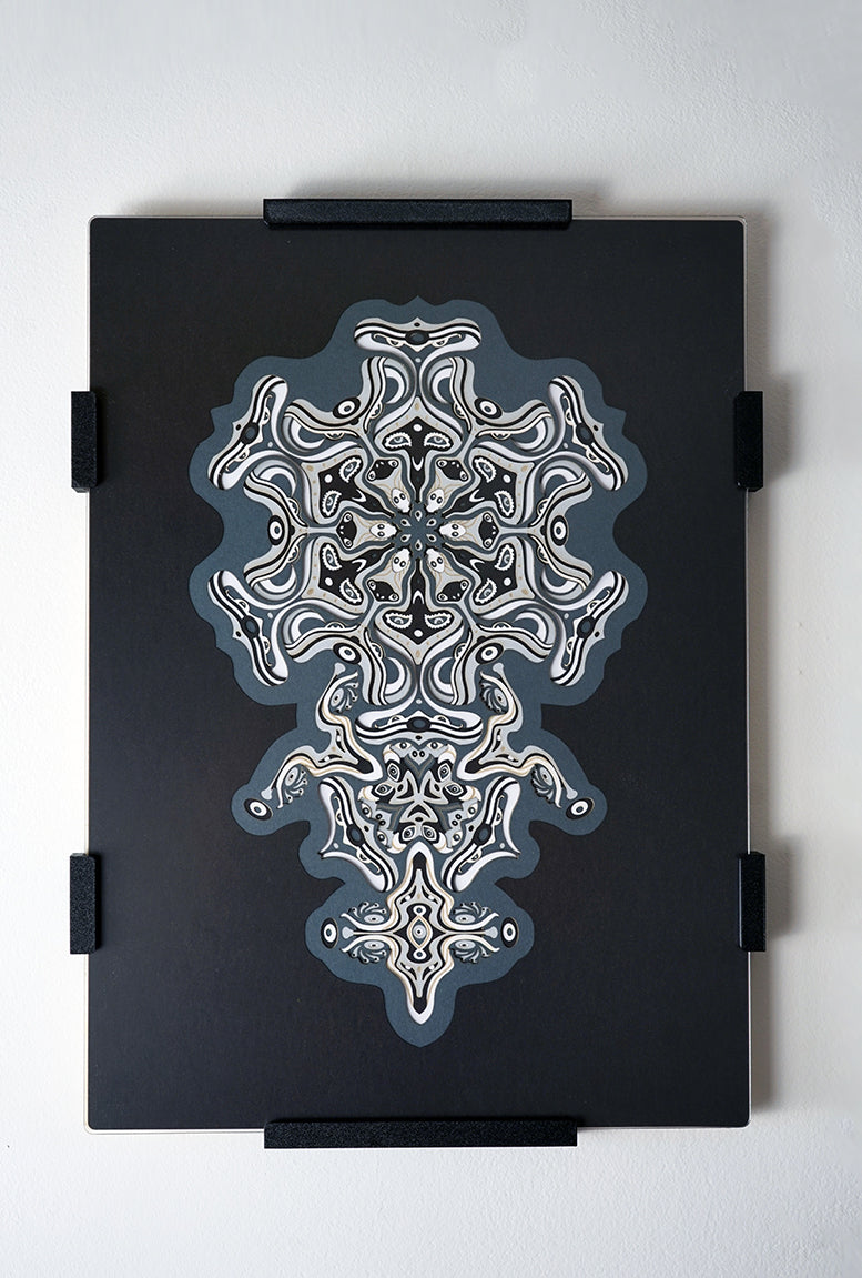 Intricate monochrome paper layers arranged in a mandala structure, creating emerging spirit faces and abstract creatures.