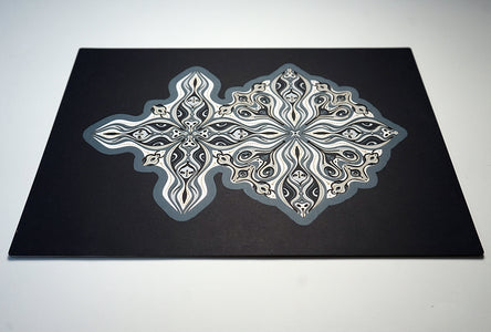 Layered grayscale paper forming symmetrical mandala patterns that reveal subtle, mask-like faces from the Metamorphs series