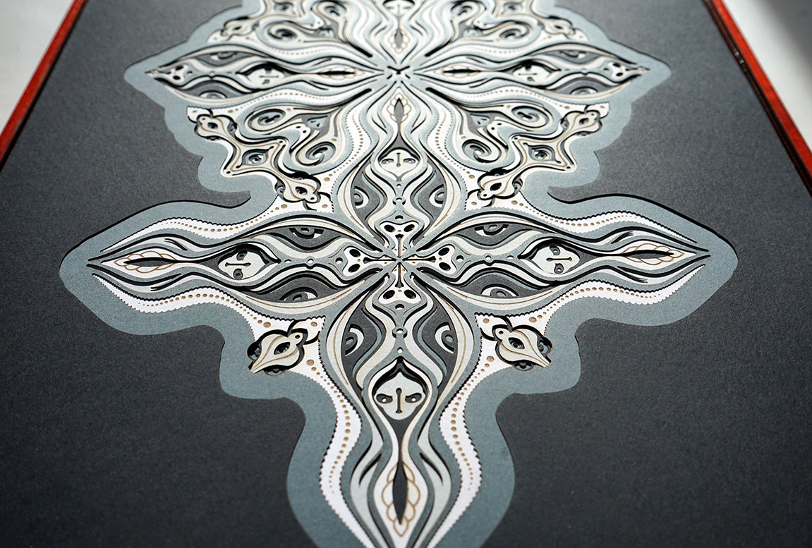Layered grayscale paper forming symmetrical mandala patterns that reveal subtle, mask-like faces from the Metamorphs series