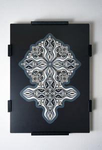 Layered grayscale paper forming symmetrical mandala patterns that reveal subtle, mask-like faces from the Metamorphs series