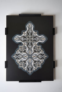 Layered grayscale paper forming symmetrical mandala patterns that reveal subtle, mask-like faces from the Metamorphs series