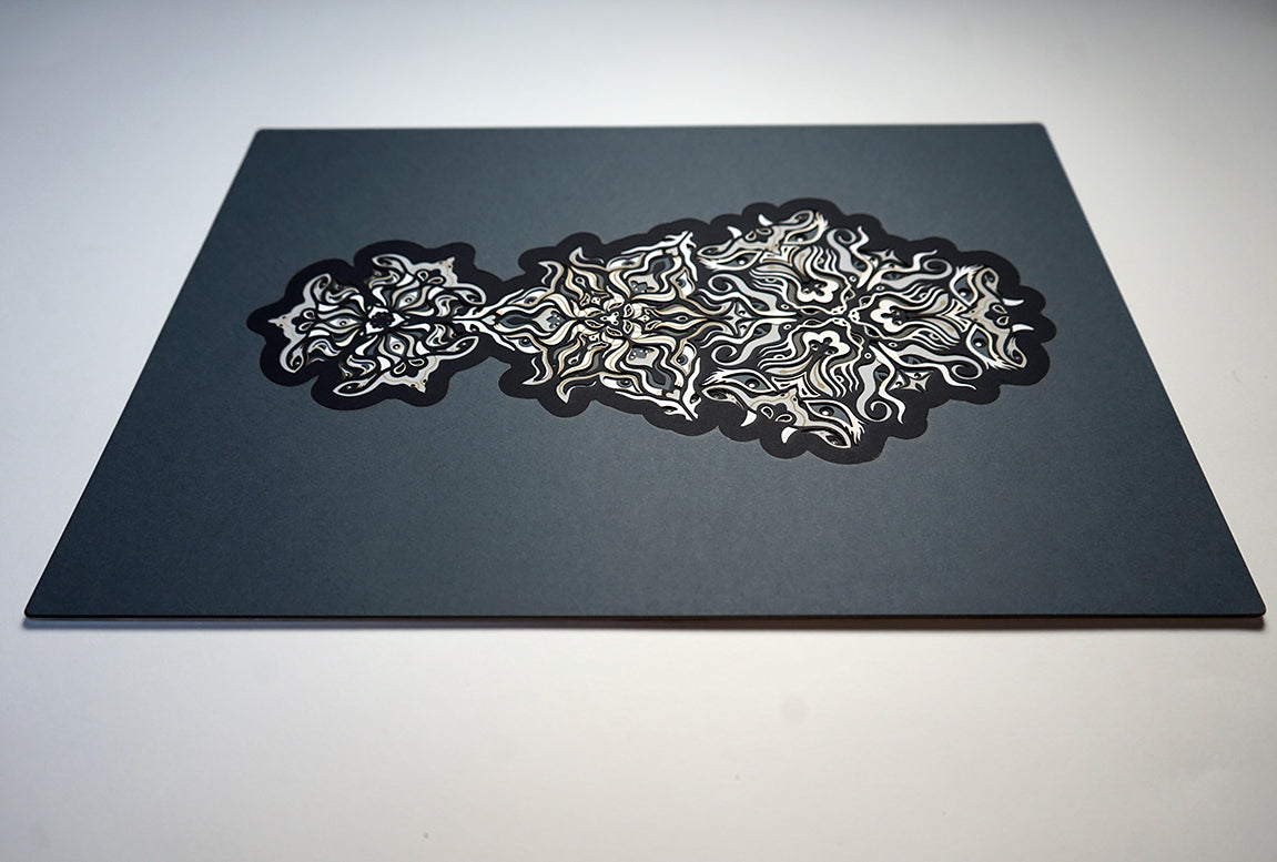 Geometric mandala made of layered grayscale paper, revealing masked spirit figures within the patterns.