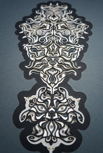 Geometric mandala made of layered grayscale paper, revealing masked spirit figures within the patterns.