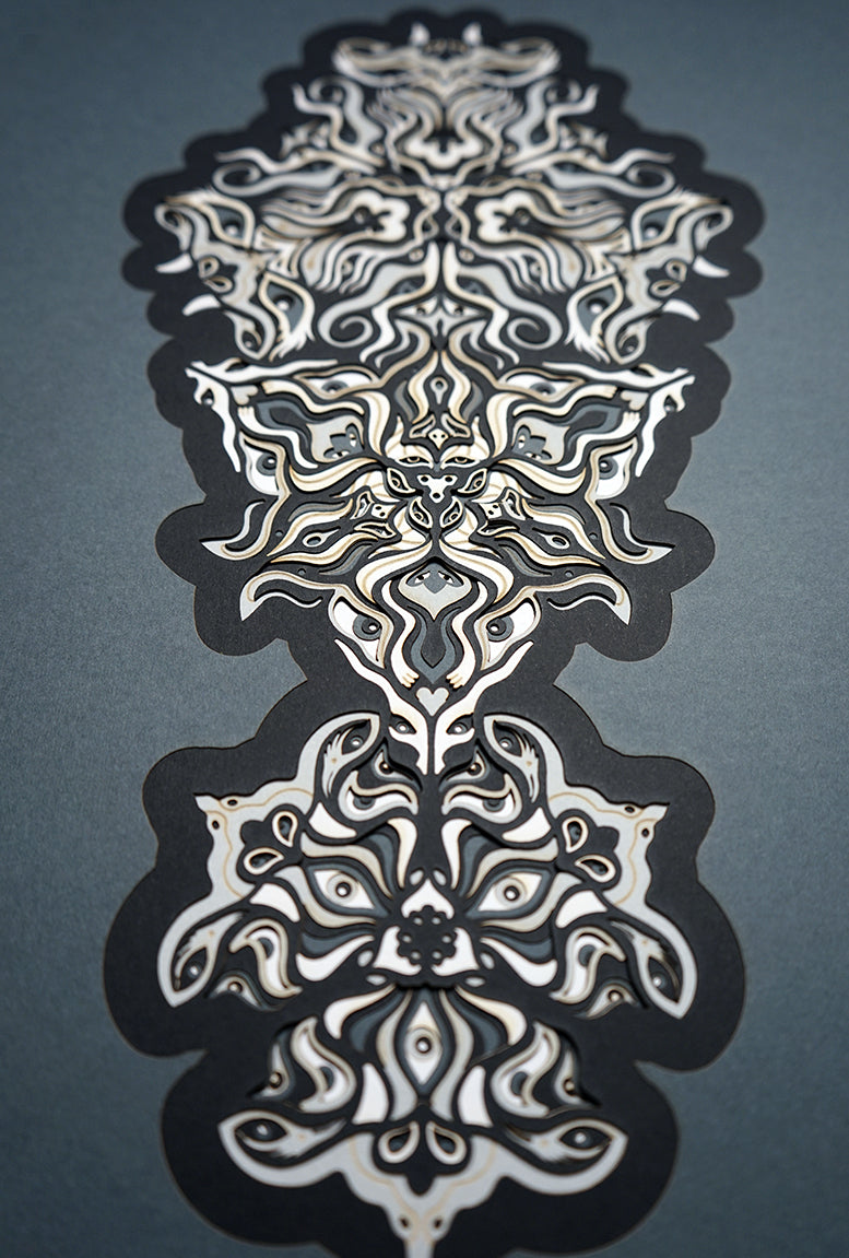 Geometric mandala made of layered grayscale paper, revealing masked spirit figures within the patterns.