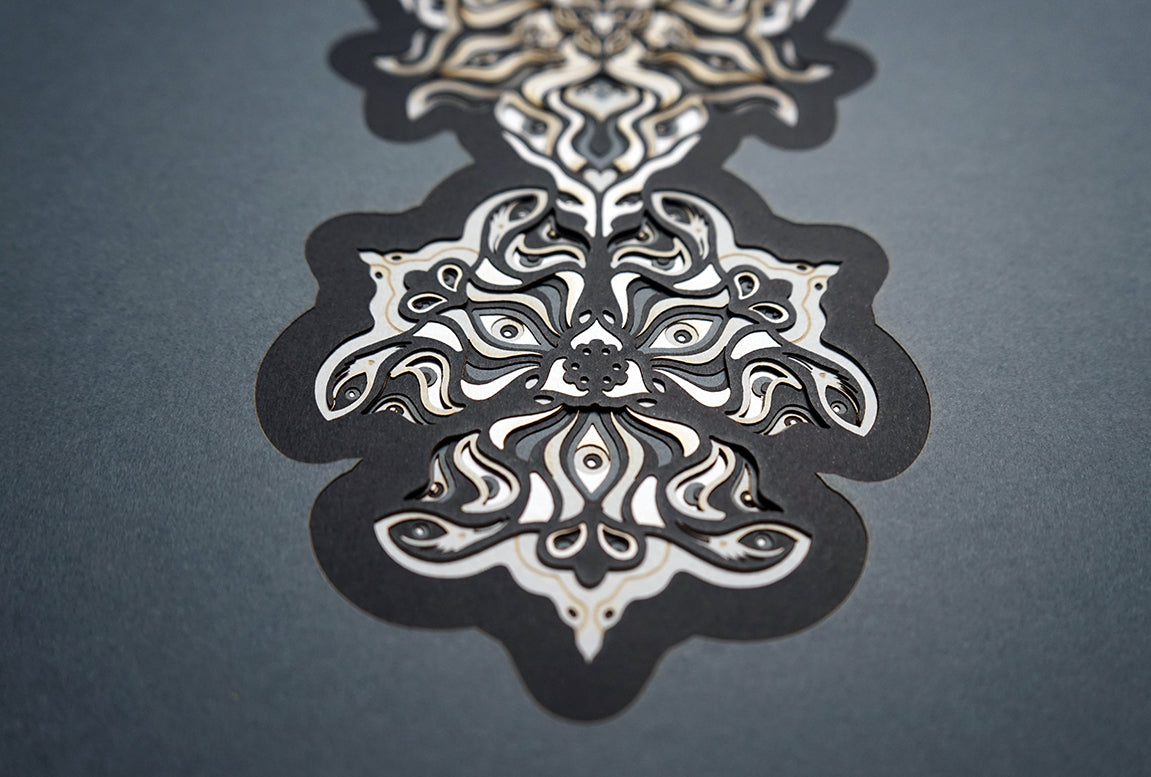 Geometric mandala made of layered grayscale paper, revealing masked spirit figures within the patterns.
