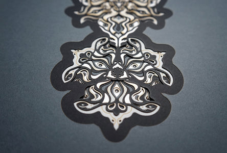 Geometric mandala made of layered grayscale paper, revealing masked spirit figures within the patterns.