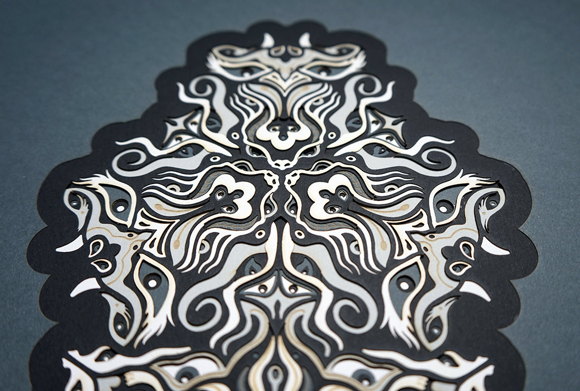 Geometric mandala made of layered grayscale paper, revealing masked spirit figures within the patterns.
