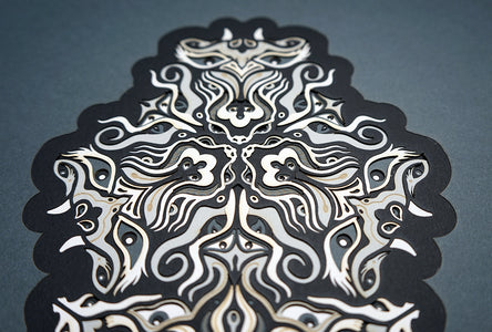 Geometric mandala made of layered grayscale paper, revealing masked spirit figures within the patterns.