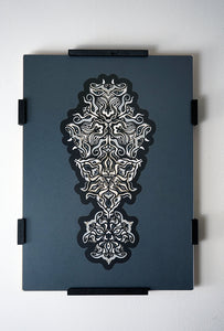 Geometric mandala made of layered grayscale paper, revealing masked spirit figures within the patterns.