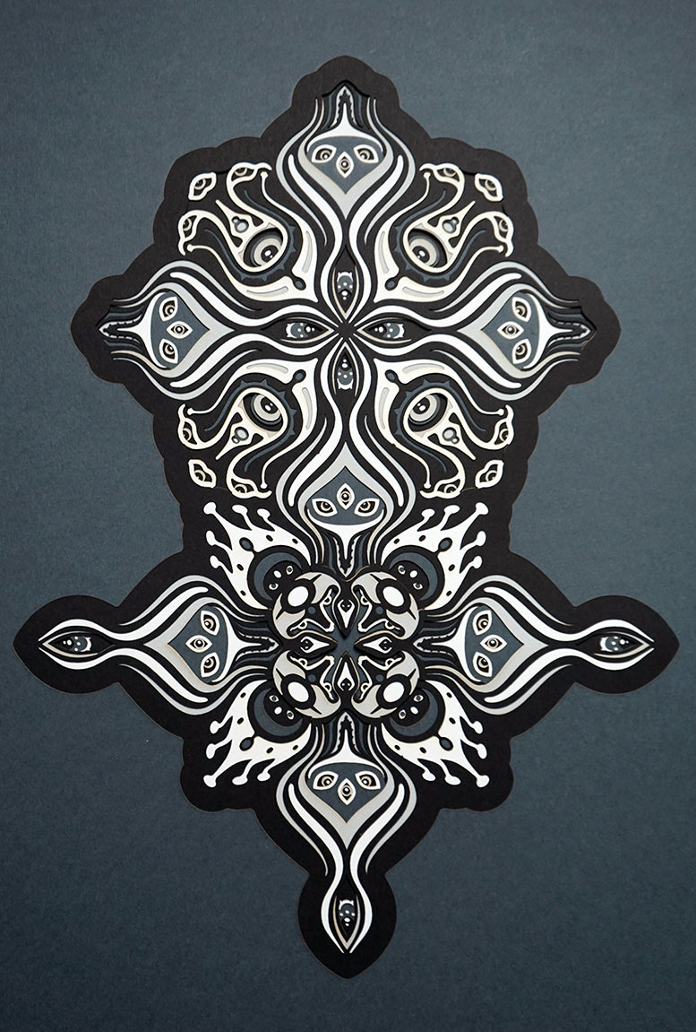 Intricate paper cut mandala in shades of black and white, with morphing faces appearing between the layers.