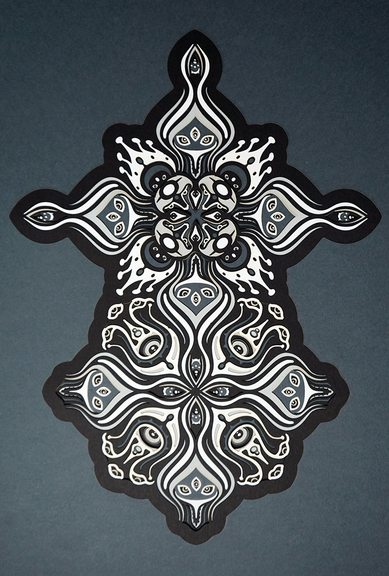 Intricate paper cut mandala in shades of black and white, with morphing faces appearing between the layers.