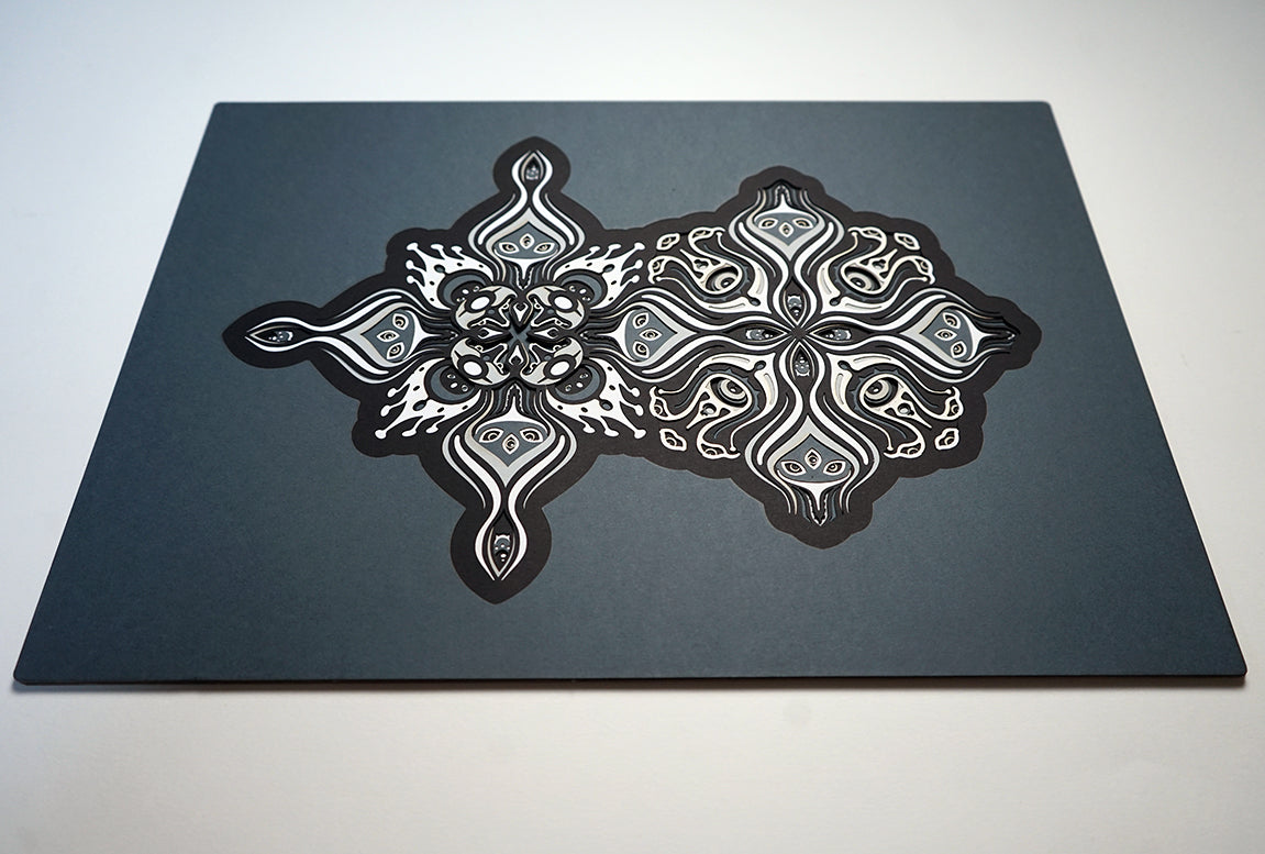 Intricate paper cut mandala in shades of black and white, with morphing faces appearing between the layers.