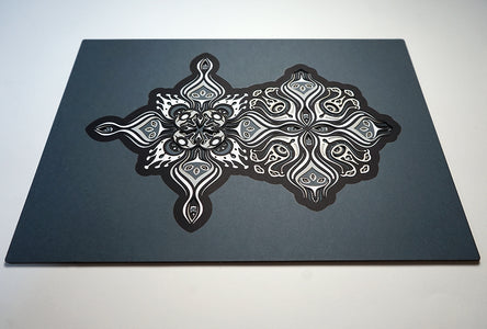 Intricate paper cut mandala in shades of black and white, with morphing faces appearing between the layers.
