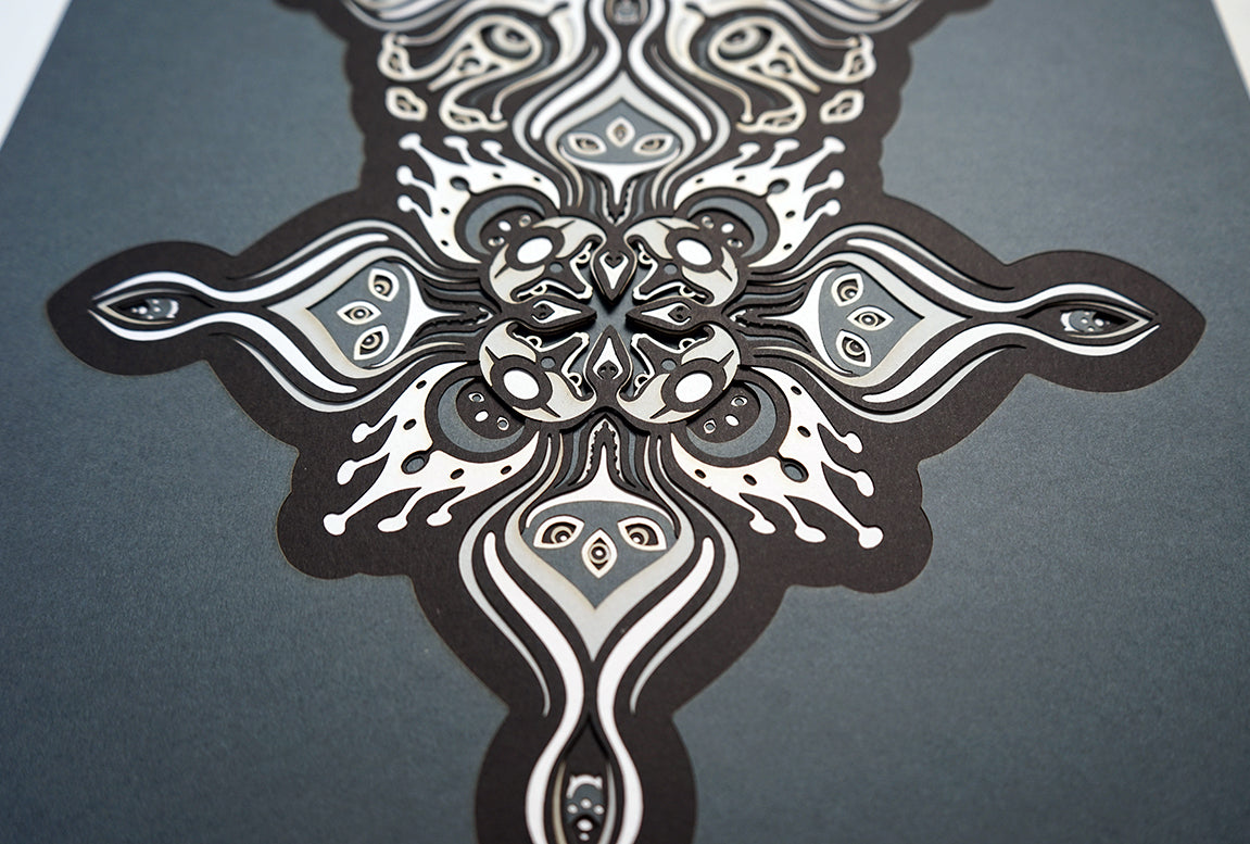 Intricate paper cut mandala in shades of black and white, with morphing faces appearing between the layers.