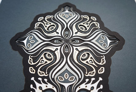 Intricate paper cut mandala in shades of black and white, with morphing faces appearing between the layers.