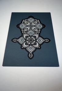 Intricate paper cut mandala in shades of black and white, with morphing faces appearing between the layers.