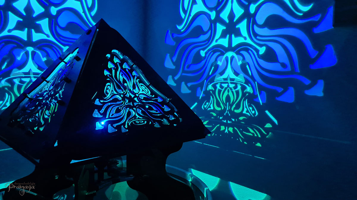 A colorful shadow lamp with a geometric design, featuring triangular sides and a pyramid shape, projecting a vibrant mushroom pattern on a wall.