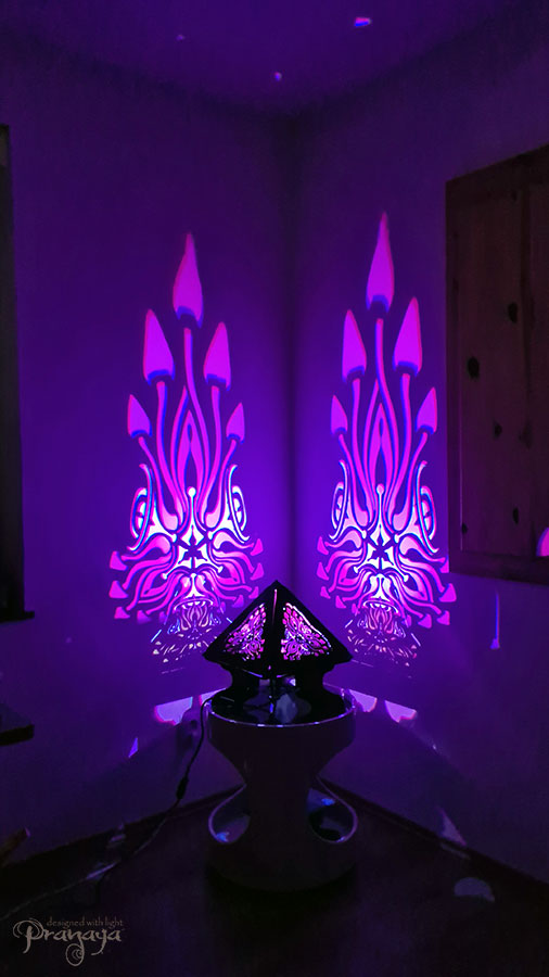 A colorful shadow lamp with a geometric design, featuring triangular sides and a pyramid shape, projecting a vibrant mushroom pattern on a wall.
