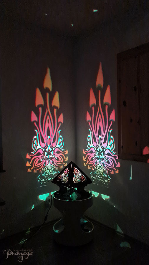 A colorful shadow lamp with a geometric design, featuring triangular sides and a pyramid shape, projecting a vibrant mushroom pattern on a wall.