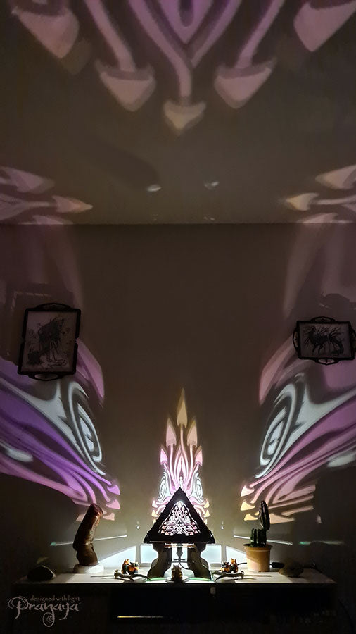 A colorful shadow lamp with a geometric design, featuring triangular sides and a pyramid shape, projecting a vibrant mushroom pattern on a wall.