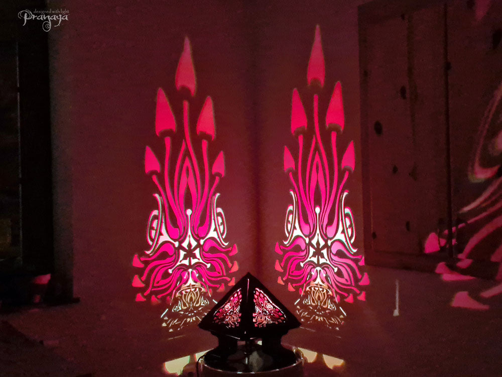 A colorful shadow lamp with a geometric design, featuring triangular sides and a pyramid shape, projecting a vibrant mushroom pattern on a wall.